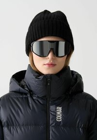 Musthave musta paksu jope kõrge kraega, külgsuunas tõmblukuga ja logoga. Must soonik kootud müts ja peegelprillid täiustavad välimust.