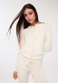 Felpa con zip color crema, con cappuccio regolabile, tasche frontali e orlo a coste. Abbinata a pantaloni della tuta color crema. Tessuto morbido.