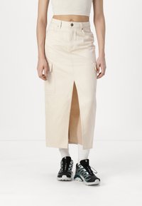 Beige midi skjørt med ett frontslitt og sidelommer, laget av bomullsmateriale. Kombinert med svarte og teal treningssko og hvite sokker.