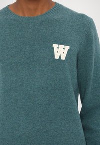 Teal knit sweater with a round neckline and ribbed cuffs. Features a white "W" logo on the chest, smooth texture.