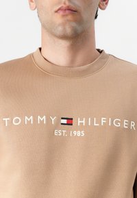 Brun sweatshirt med rund hals, der har et hvidbroderet "TOMMY HILFIGER" logo og en lille detalje med rød, hvid og blå flag.