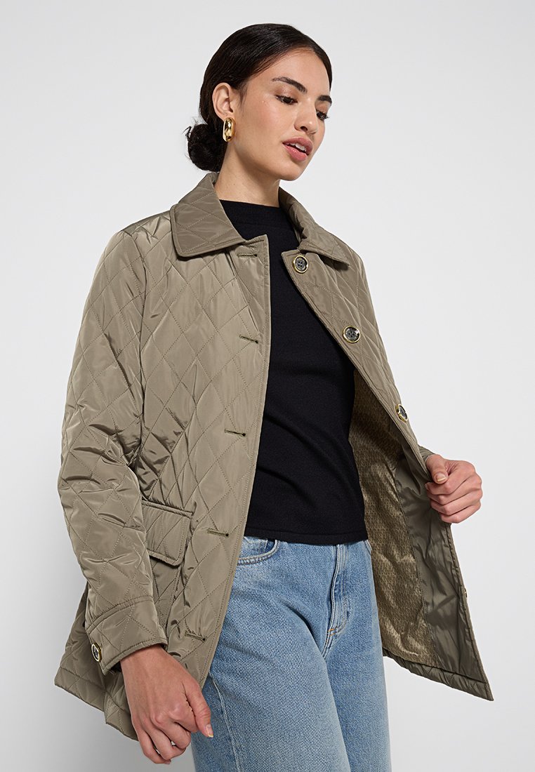 Beige gesteppte Jacke mit Kragen, vorderen Knöpfen und zwei Seitentaschen. Innen mit einem strukturierten Futter ausgestattet. Getragen über einem schwarzen Oberteil.