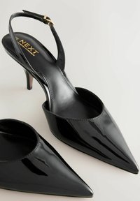 Fekete lakkbőr pántos magassarkú cipő, hegyes orral, finom varrással és vékony sarokkal. Aranyszínű csatt díszíti a pántot.