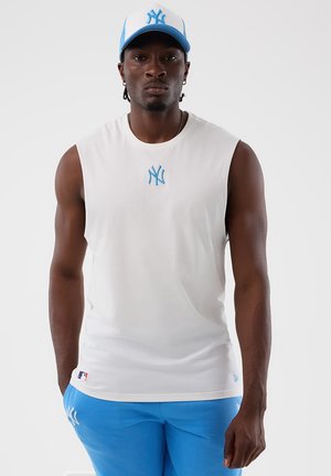 Uomo che indossa una maglietta bianca senza maniche e un cappellino blu con il logo dei New York Yankees, in piedi davanti a uno sfondo bianco semplice.