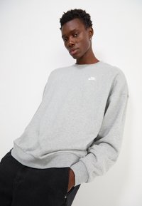 Sweatshirt cinza feito de um tecido suave e texturizado, apresentando um logo branco da Nike no peito e punhos canelados. Combinado com calças pretas.