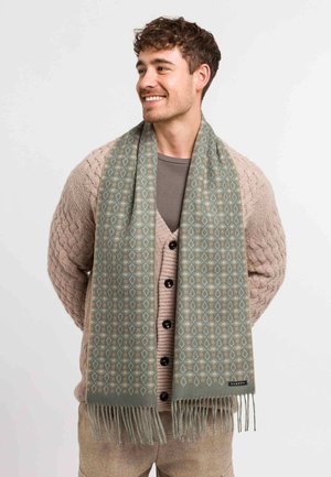 Helles grünes Schal mit geometrischem Muster, fransigen Kanten und weicher Textur, getragen über einem beigen Strickcardigan und einem grauen Hemd.