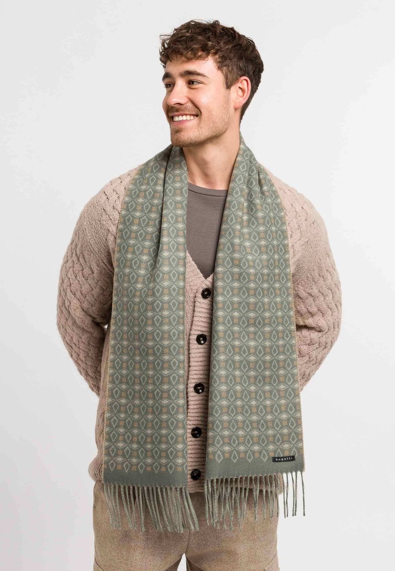 Ljusgrön scarf med ett geometriskt mönster, fransade kanter och en mjuk textur, bärs över en beige stickad kofta och en grå skjorta.