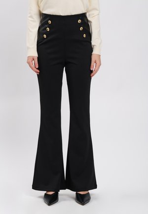 Schwarze, hoch taillierte ausgestellte Hose mit sechs goldenen Knöpfen und schwarzen spitzen Schuhen, getragen zu einem cremefarbenen Langarmshirt.