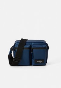 Blue messenger bag with two front zippered pockets, adjustable strap, and textured fabric. Features a logo patch and black accents.