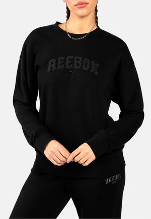 Frau trägt schwarzen Reebok-Pullover und Hose, mit strukturiertem Reebok-Logo auf der Vorderseite des Pullovers und der Hose, Hände unterhalb der Taille verschränkt.