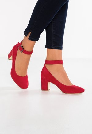 Czerwone zamszowe szpilki z okrągłym noskiem i paskiem na kostkę, z klamrą. Obcas ma kształt bloku z jasnym akcentem.