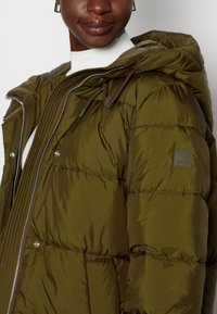 Chaqueta de plumas verde oliva con capucha, cierre frontal con cremallera y botones a presión; cuenta con ribetes elásticos y detalles de parches en la manga.