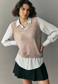 Beżowy sweter bez rękawów z dekoltem w serek, nałożony na białą koszulę w czarne paski, w połączeniu z czarną plisowaną spódnicą.