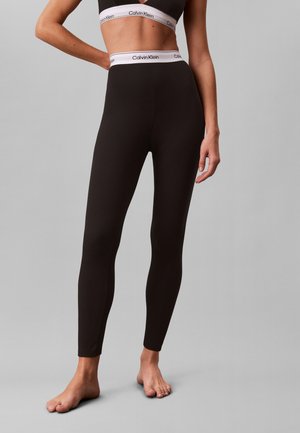 Kvinde iført sorte højtaljede leggings og matchende sort sports-bh med hvide Calvin Klein elastikker, stående barfodet på gråt gulv.