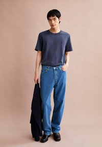 Navyblauwe t-shirt met een klein logo, gecombineerd met ruim zittende blauwe jeans en zwarte klompen. Donkere jas over één arm gedrapeerd.