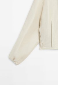 Lange beige linnenjas met mouw met elastische manchetten, zichtbare zijnaad en subtiele textuur op een witte achtergrond.