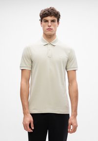 Polo shirt beige clair avec col, deux boutons au cou et des finitions contrastantes sur les manches. Présente un petit logo emblématique.
