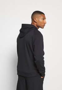 Sudadera negra hecha de tejido suave, con un bolsillo canguro, puños de canalé y capucha con cordón, mostrada de lado con un acabado liso.