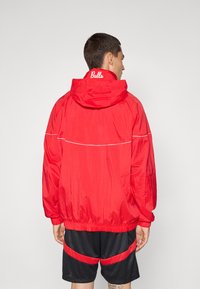 Rød hættejakke med en skinnende tekstur, indsnævret talje og hvid accentlinie. "Bulls" logo på ryggen. Bæret med sorte shorts.
