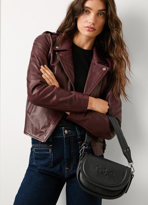 Black leather crossbody bag with a curved design and embossed logo. Model wearing a burgundy leather jacket and dark denim jeans.