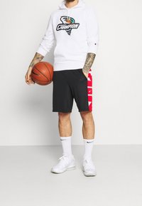 Vit hoodie med "CHAMPION" grafik, svarta shorts med röda sidopaneler som har Nike-loggor, vita strumpor och vita sportskor, håller i en basketboll.