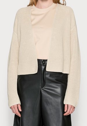 Cardigan beige a maglia con davanti aperto, caratterizzato da una texture a coste e maniche lunghe. Abbinato a pantaloni neri in pelle a vita alta.