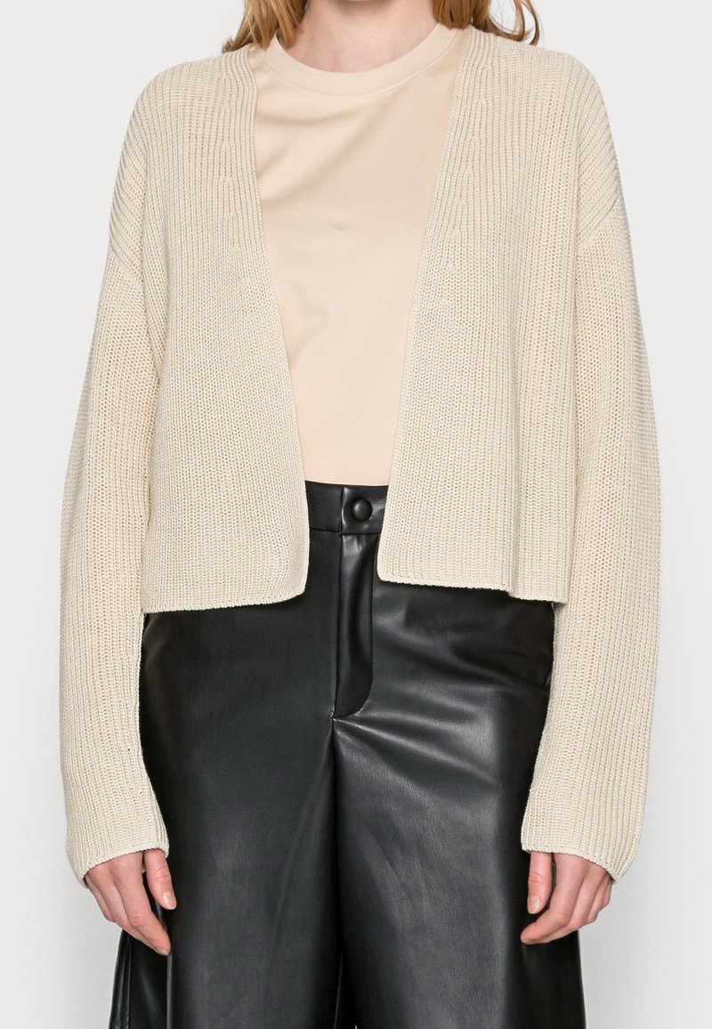 Beige Strickjacke mit offenem Vorderteil, ausgestattet mit gerippter Textur und langen Ärmeln. Kombiniert mit schwarzen, hoch taillierten Lederhosen.