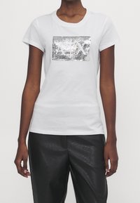 Biała bawełniana koszulka z okrągłym dekoltem i krótkimi rękawami, z prostokątnym wzorem z srebrnych cekinów na przodzie. W zestawieniu z czarnymi błyszczącymi spodniami.