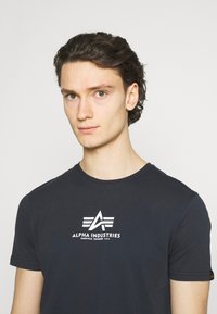 Mørkeblå bomull T-skjorte med rund hals, kortermet og med et hvitt logodesign på brystet som har teksten "ALPHA INDUSTRIES."