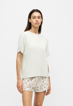 Kvinde iført en simpel hvid T-shirt med korte ærmer og hvide shorts med et lyserødt floral mønster, stående mod en ensfarvet hvid baggrund.