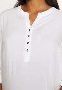 Bílá košile na knoflíky s légou obsahující pěti černými knoflíky, kulatým výstřihem a třičtvrtinovými rukávy vyrobená z lehkého materiálu.