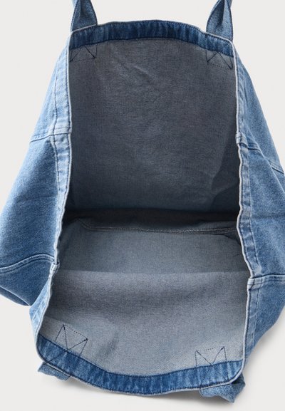 Leeg blauwe denim draagtas met brede hengsels, met zicht op de binnenstof en stiksel details op een lichte achtergrond.
