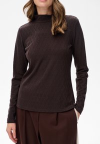 Frau in einem dunkelbraunen gemusterten Langarm-Turtleneck und lockeren braunen Hosen, posiert mit einer Hand in der Tasche.