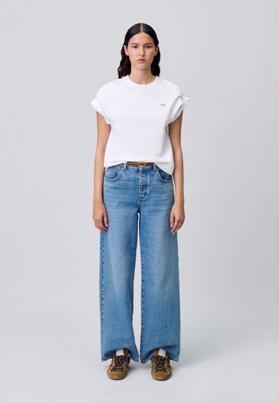 T-shirt bianco in cotone con maniche arrotolate, abbinato a jeans ampi blu chiaro. Sneakers marroni con lacci completano l'outfit.