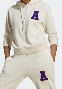 Krämfärgad hoodie och matchande sweatpants i mjukt material. Har broderat rött bokstav 'A' och lodrätt randdetalj på sidan.