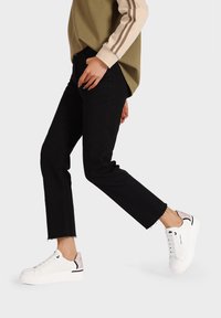 Jeans neri a vita alta abbinati a sneakers bianche con un accento rosa sul tacco. L'outfit è completato da un top a maniche lunghe marrone e beige.