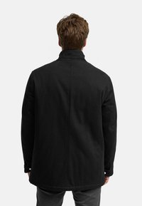 Svart frakk med høy krage, teksturert stoff og rett snitt, med minimalistisk design og knappdetaljer på ermene.