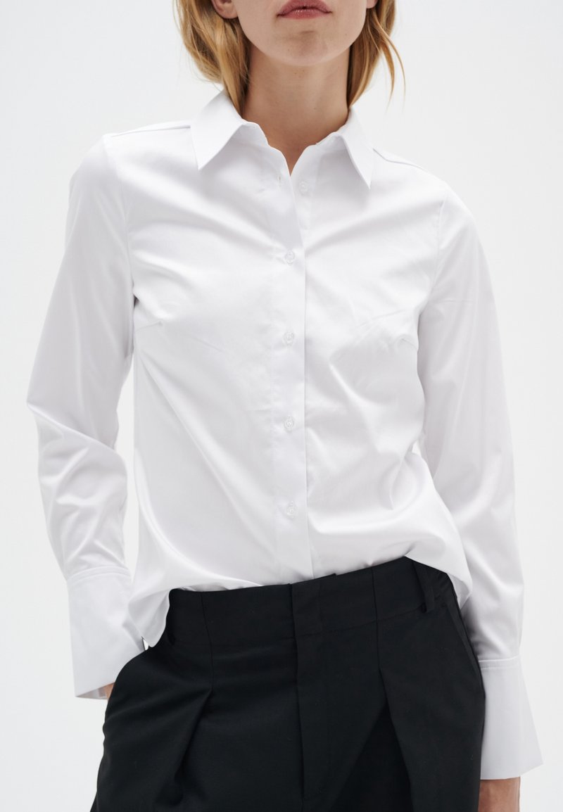 Femme portant une chemise blanche ajustée à boutons avec de longs poignets et un pantalon noir plissé, mains partiellement dans les poches.