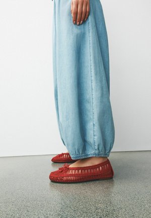 Persoon die lichtblauwe broek met elastische zoom en rode geweven instapschoenen draagt, staand op een grijs gepolijste vloer.