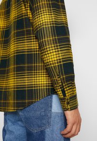 Geel-groen geruit overhemd met lange mouwen en knoopmanchet. De stof lijkt geweven en heeft een zachte textuur. Draagt over een blauwe patchwork spijkerbroek.