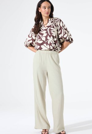 Bruine en witte blouse met bloemmotief en opgeblazen mouwen, gecombineerd met lichtbeige wijde broeken. Model staat met handen op de heupen.