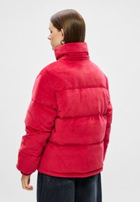 Sarkans pūka mētelis ar augstu apkakli, mīkstu tekstūru, kvadrātveida dizainu un elastīgām piedurknēm, skatīts no aizmugures.