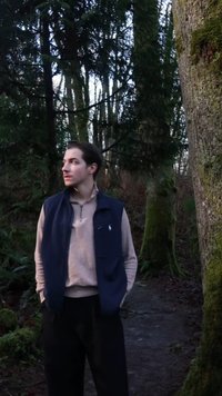 Een marineblauw vest over een lichtbeige trui, gecombineerd met een donkere broek. Staand tegen een met mos bedekte boom in een bosrijke omgeving.