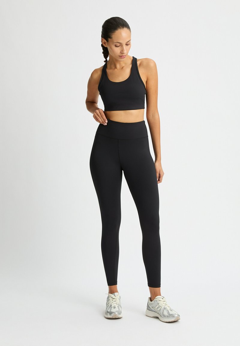 Un reggiseno sportivo nero e leggings a vita alta, entrambi in un materiale liscio e elastico. Le sneakers con un design grigio strutturato completano l'outfit.