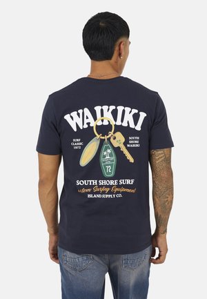 Man met getatoeëerde arm die een marineblauw Waikiki surf-thema T-shirt draagt met een sleutelhangerafbeelding en tekst over South Shore Surf.