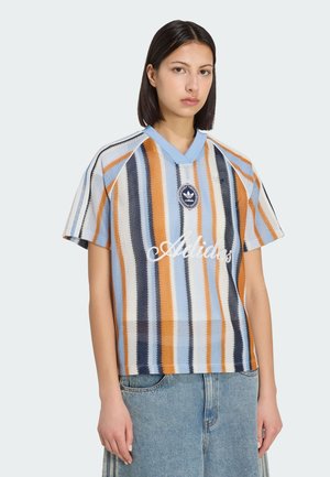 Kobieta o ciemnych włosach, ubrana w prążkowaną dzianinową koszulkę Adidas w kolorach niebieskim, pomarańczowym, białym i czarnym, połączoną z jasnoniebieskimi jeansami.