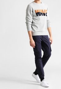 Felpa grigia con la scritta "WAVE" in arancione e blu navy, abbinata a pantaloni cargo blu navy e sneakers bianche. Presenta polsini a costine e contrasto di texture.
