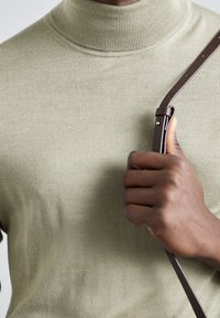 Zelený pletený svetr s rolákem a hladkou texturou, s přiléhavým střihem. Osoba drží hnědý kožený popruh s kovovou sponou.