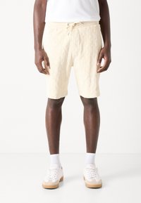 Cream-colored shorts with a diamond-patterned texture, elastic waistband, drawstring detail, and knee-length cut. Paired with white sneakers.