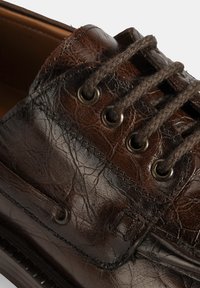 Braune Lederschuhe mit einer strukturierten Oberfläche, ausgestattet mit Metallösen, braunen Schnürsenkeln und Lederverzierungen entlang der Kanten.